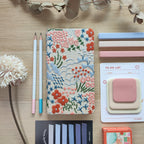 Quaderno Dery, copertina rigida con fiori ricamati, su scrivania in legno con matite post-it puzzle fiore, BECCA